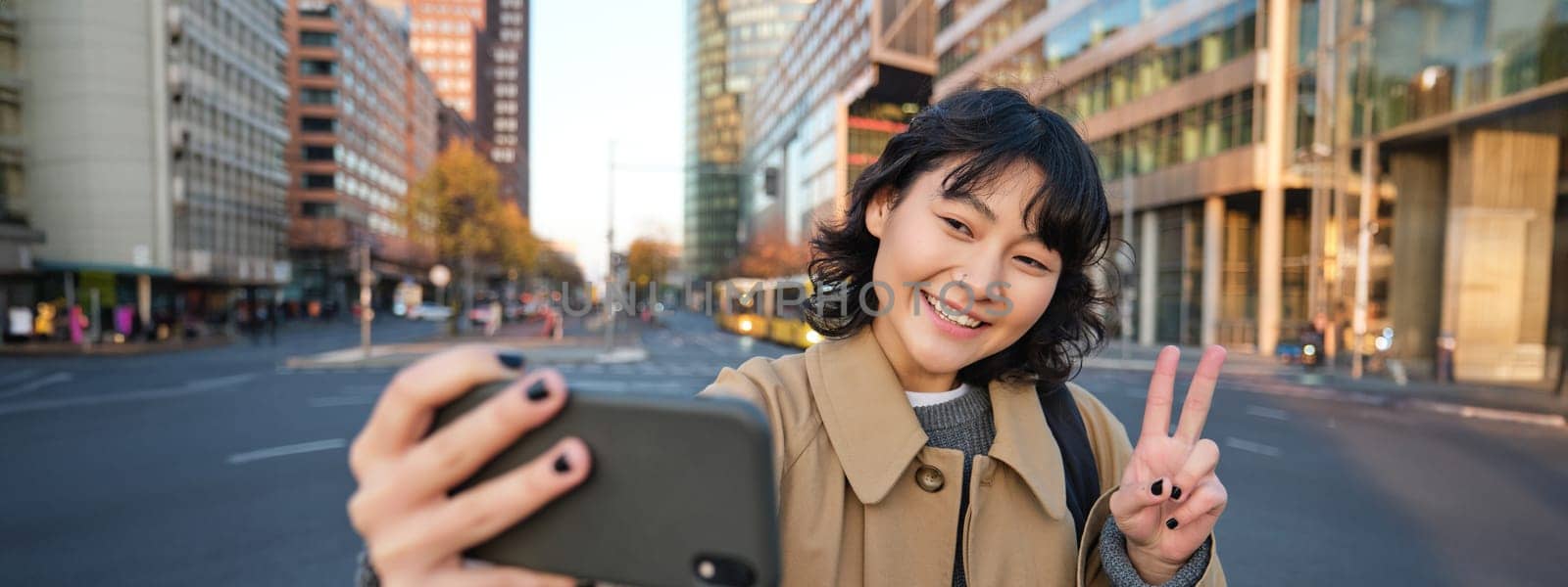 Happy asian girl, tourist takes photo in city centre, shows peace sign at smartphone camera, makes selfie during sightseeing with smiling joyful face by Benzoix