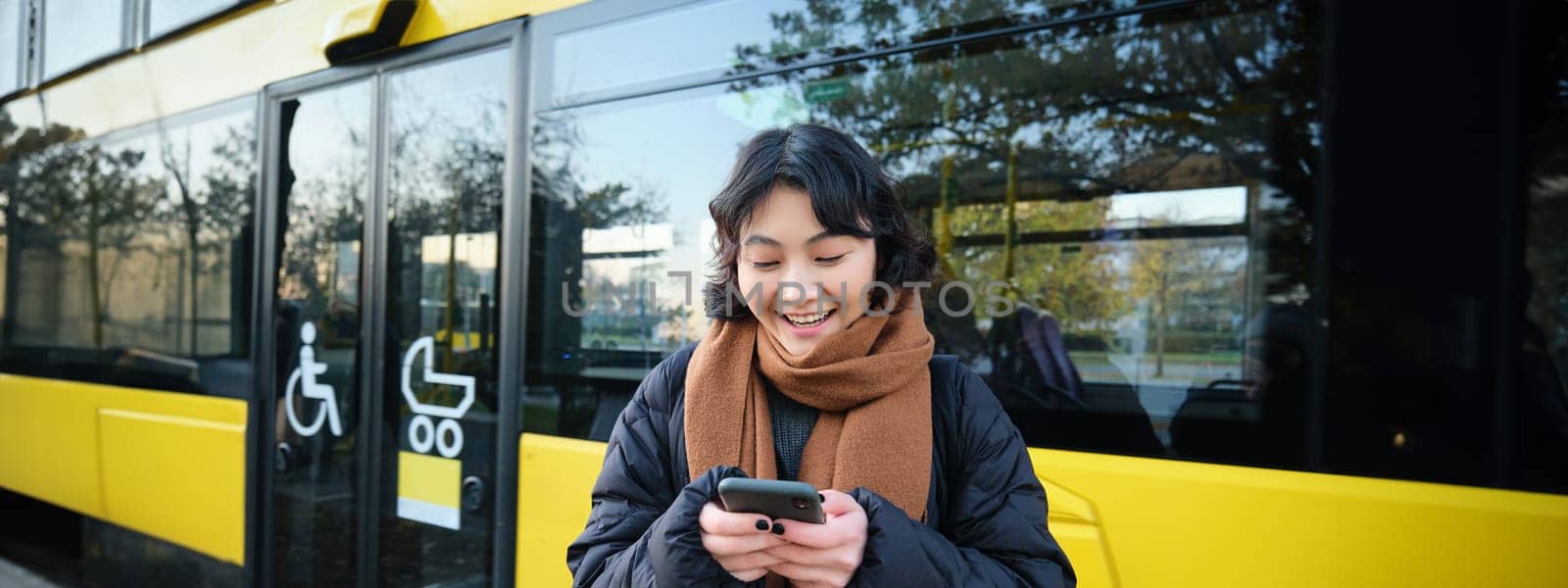 Beautiful korean girl, student on bus stop, looking at her smartphone, checking timetable, reading text message, wearing winter clothes by Benzoix