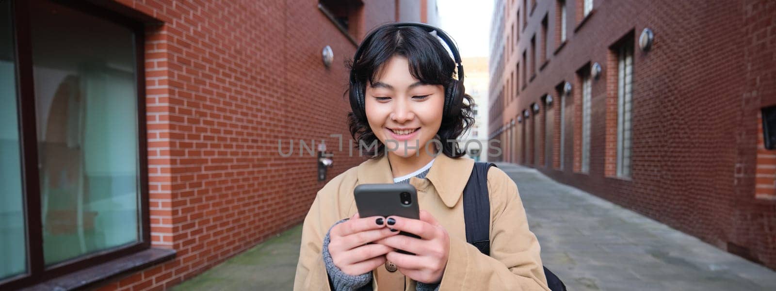 Portrait of young korean woman walking around city with backpack and headphones, listens music, looks at smartphone, uses phone application on streets by Benzoix