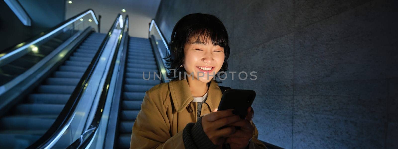 Smiling korean girl going down escalator in dark, holding mobile phone, using smartphone app, listening music, commuting in city by Benzoix