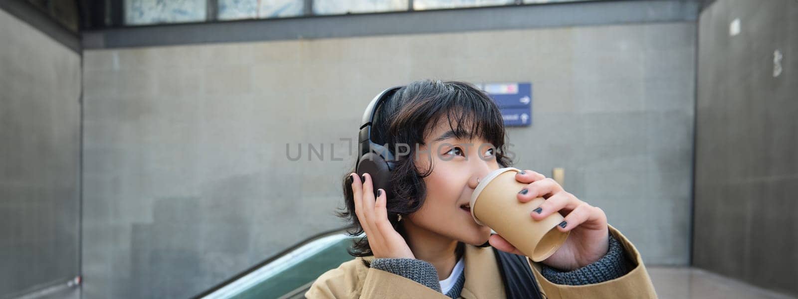 Portrait of stylish asian woman in headphones, drinks coffee to go and smiles, enjoys cappuccino while commutes, stands on street by Benzoix