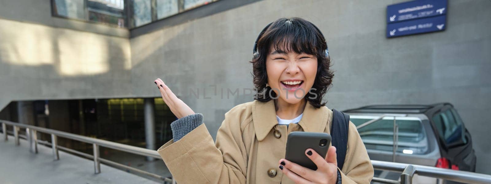 Happy asian girl in headphones, looking at mobile phone and celebrating, reading message with good news, screams yes with excitement, stands on street.