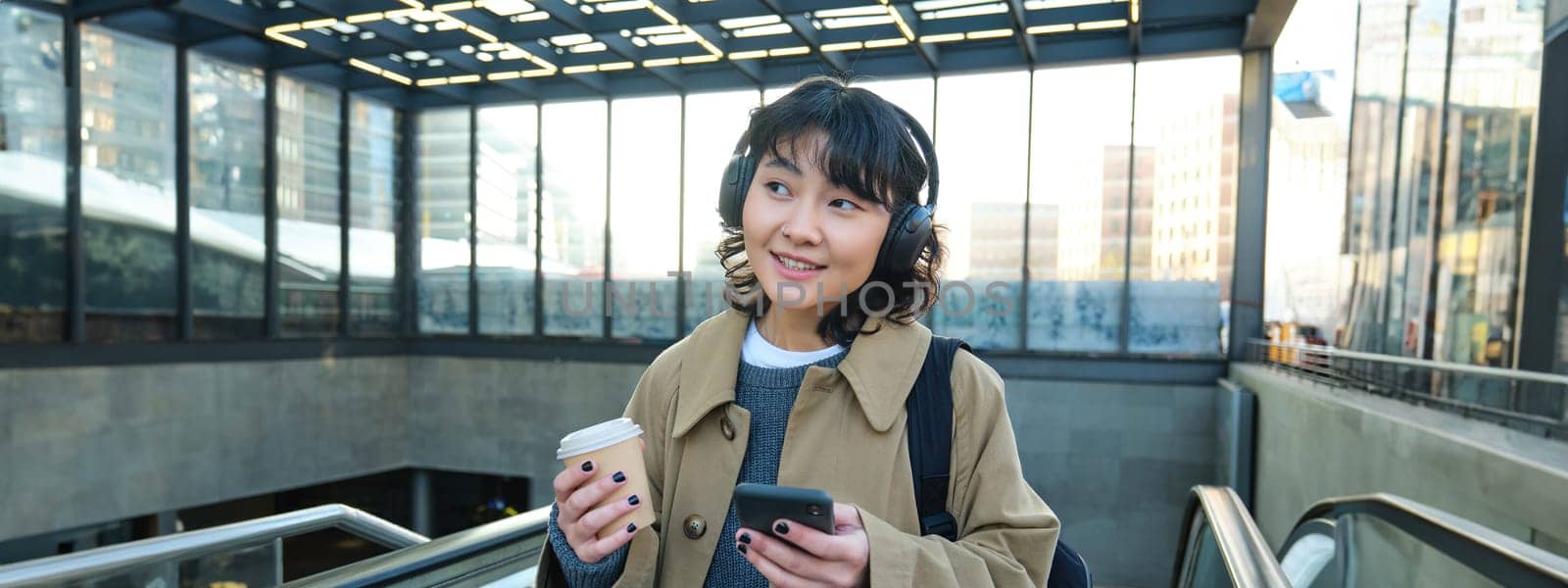 Portrait of smiling korean girl drinks coffee to go, goes up an escalator, holds smartphone, visits new city, arrives at train station, listens music in headphones.