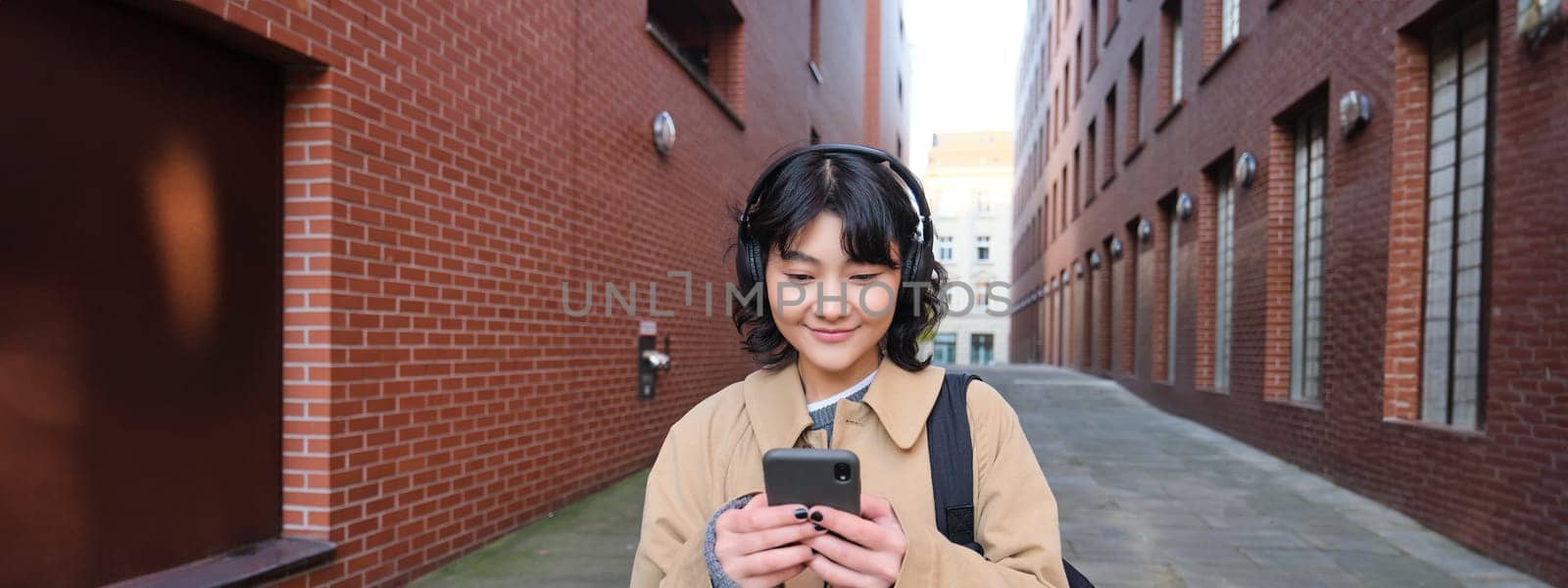 Young asian woman listens music in wireless earphones, uses smartphone on street, walks around city on sunny day, smiling happily by Benzoix