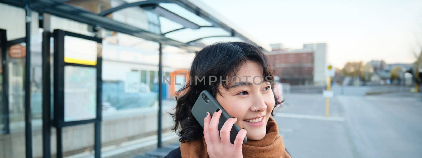 Close up of young asian woman talks on mobile phone, has conversation over telephone, waits for bus on public transport stop.