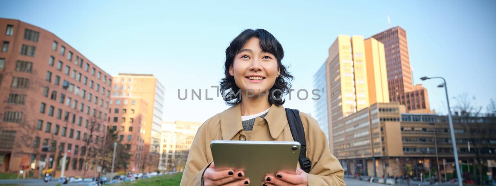 Happy asian girl stands on street, university student walks with digital tablet in hands and smiles, stands in city centre.