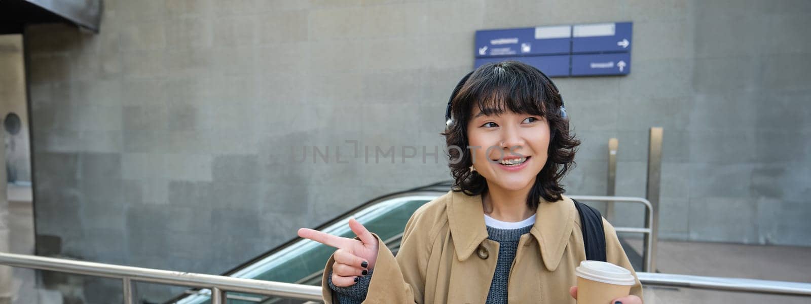 Happy brunette woman in headphones, listens music, enjoy drinking cup of coffee, stands on street, train station.