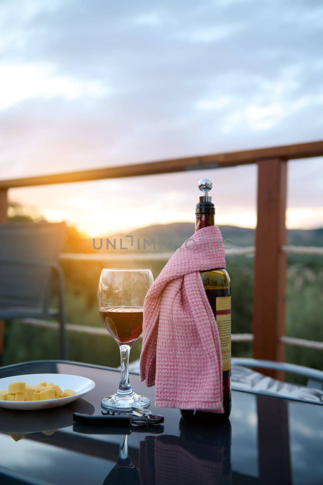 Glass of wine with delicious red wine near a plate with sliced cheese on a summer terrace against sunset background. Romantic dinner. Aperitif. Food background