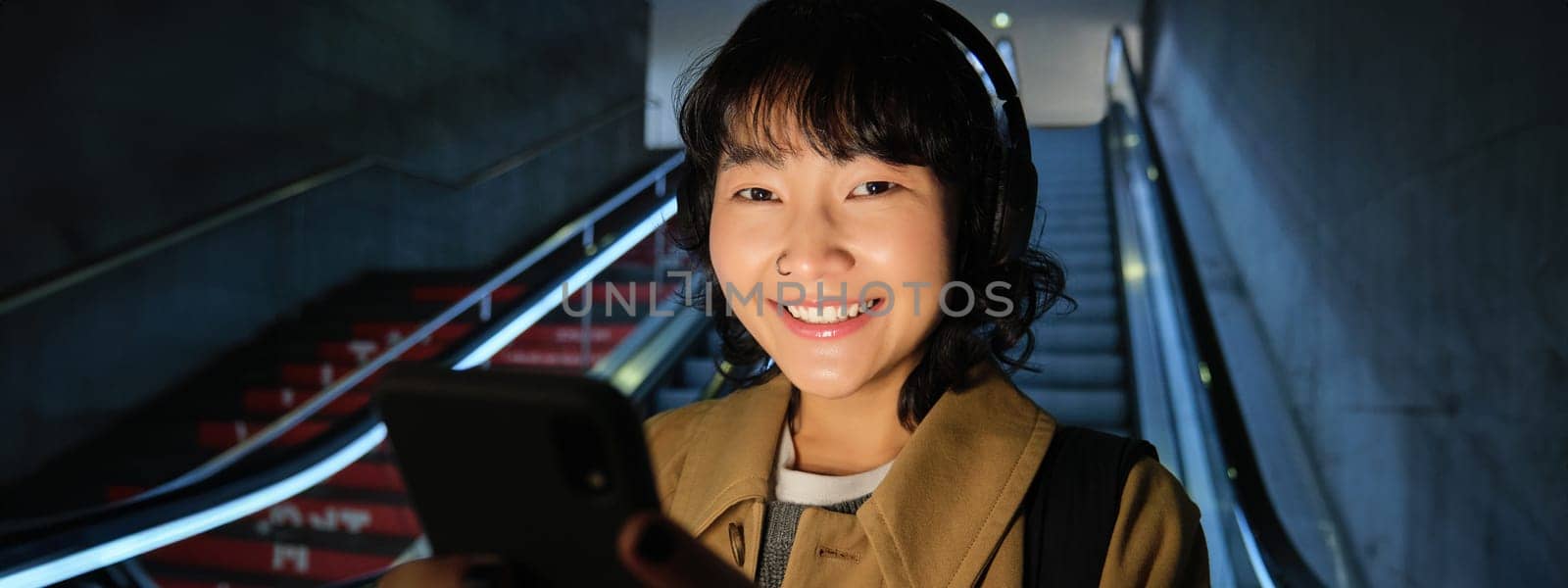 Close up of cute asian girl in headphones, picks song to listen while commuting, going down escalator to metro, smiling as reading message on mobile phone.