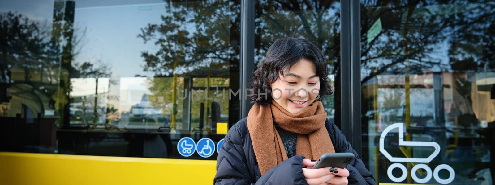Young beautiful woman standing on bus stop, texting message on smartphone, holding mobile phone, checking her schedule, buying ticket online, wearing winter clothes.