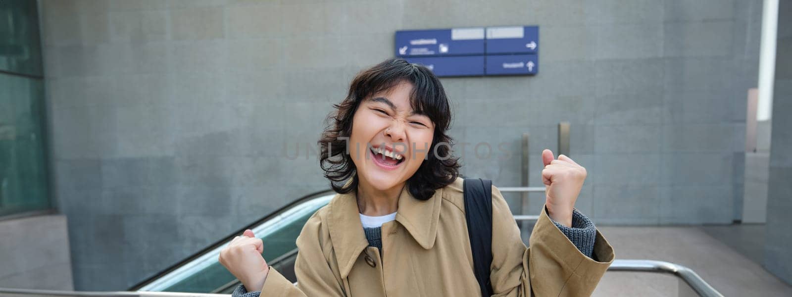 Enthusiastic asian girl stands on street, makes hooray celebration gesture, says yes, triumphing, winning and enjoying this feeling, standing alone outdoors by Benzoix