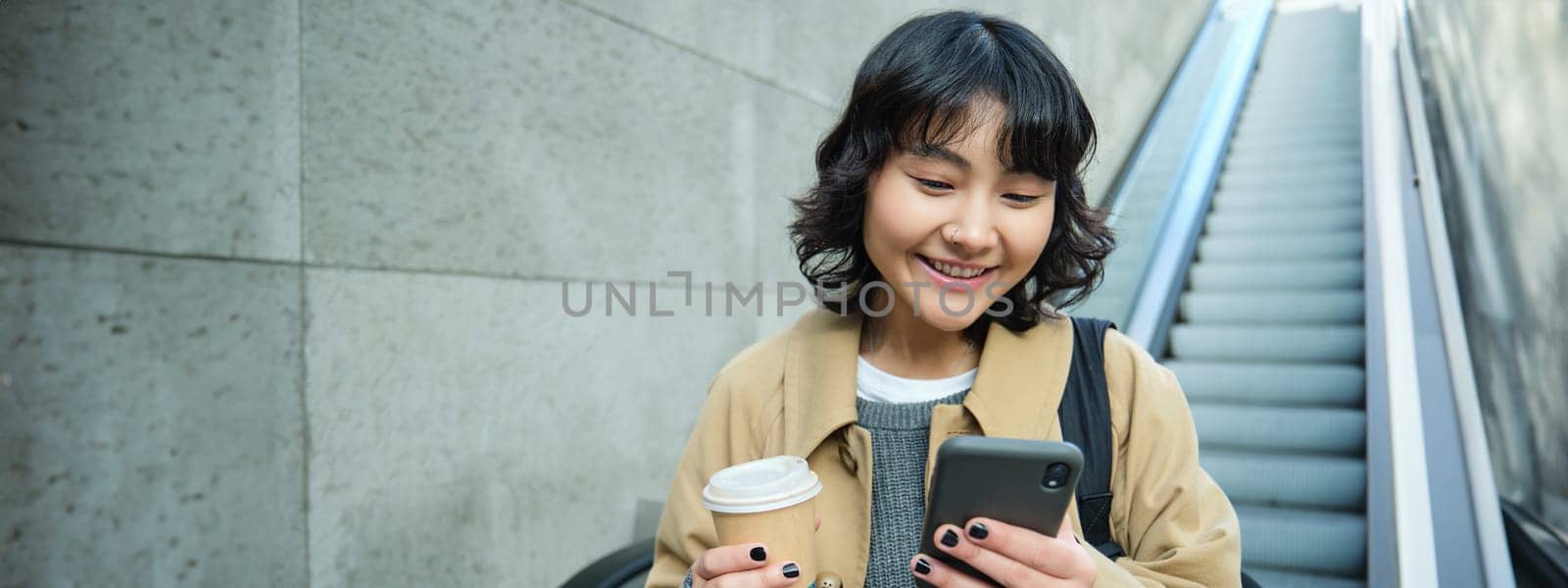 Portrait of smiling korean girl commutes, goes somewhere in city, drinks coffee to go and uses smartphone, stands on escalator by Benzoix