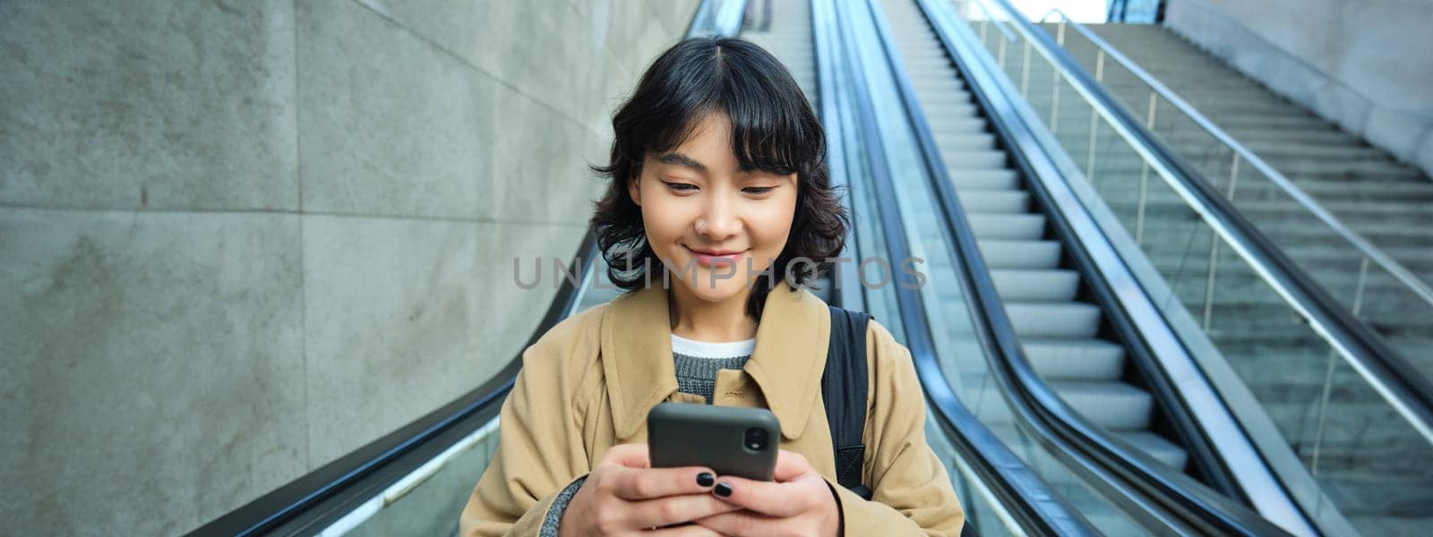 Portrait of beautiful asian girl, student goes down escalator in city, looks at mobile phone, uses telephone, map application, commutes somewhere in town.
