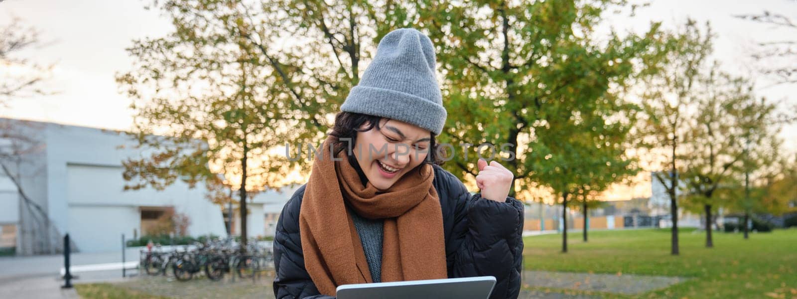 Portrait of happy asian girl sits on bench, looks at digital tablet screen and cheers, triumphs, wins something, celebrates good news, relaxes in park outdoors.