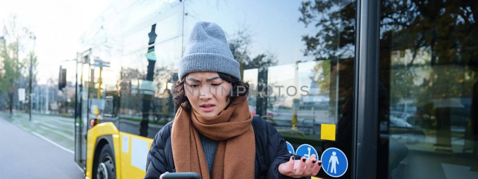 Portrait of confused young woman, looking at her smartphone app with shocked and disappointed face, shrugs shoulders, reads bad news, stands on bus stop near public transport by Benzoix