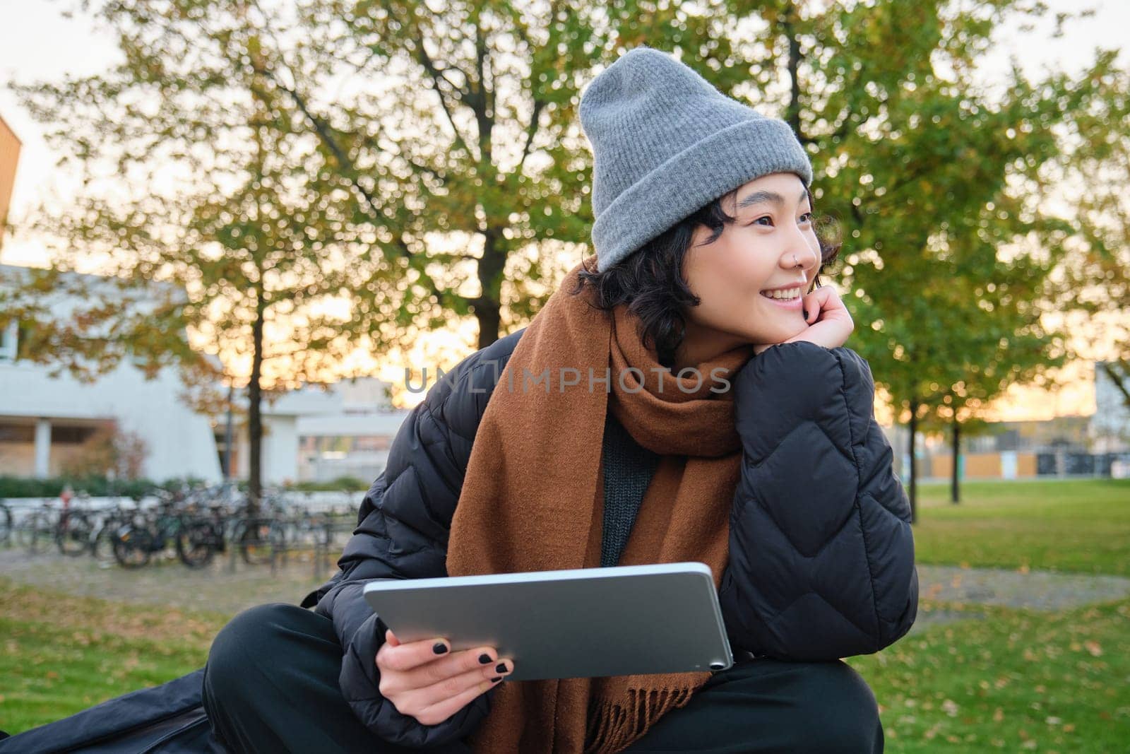 Smiling asian girl, student sits on bench in park alone, reading, using social media app on digital tablet, watches videos outdoors, relaxes on fresh air.