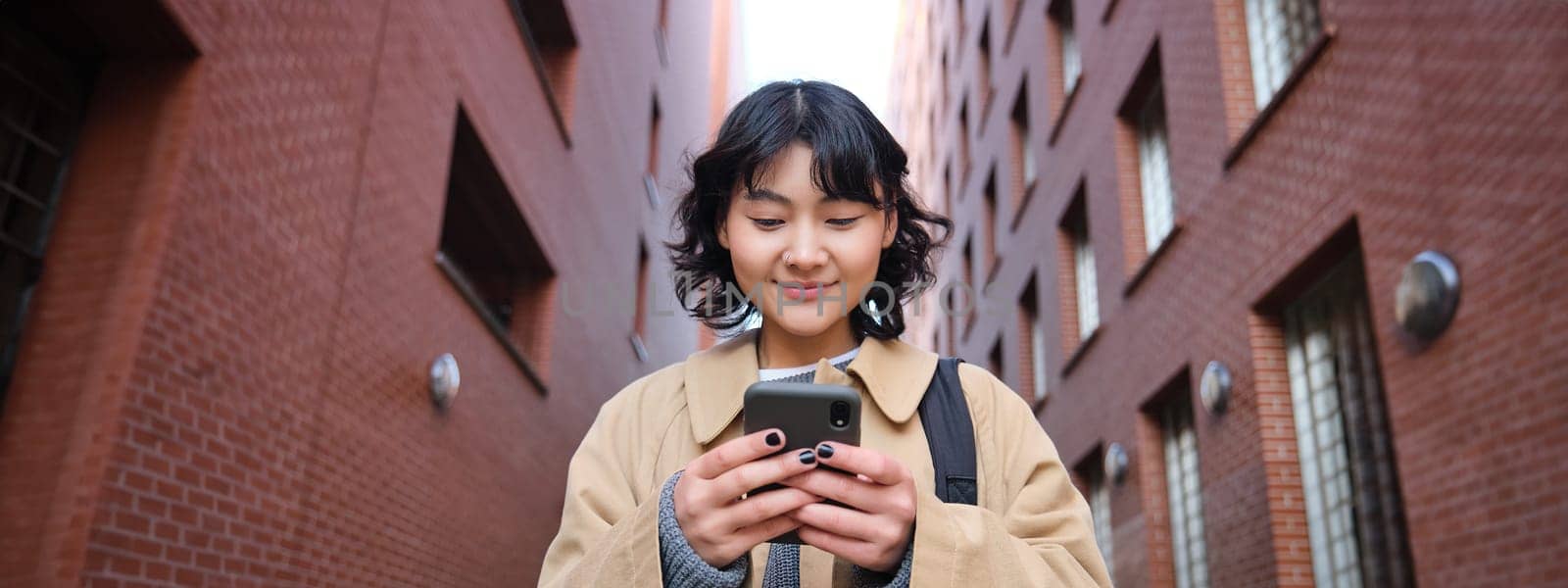 Lower angle view of brunette korean girl, listening music in headphones, walking along street and looking at smartphone, reading message on mobile phone by Benzoix