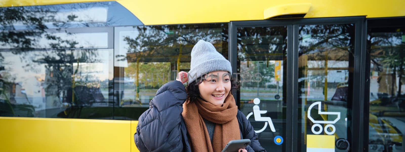 Portrait of girl standing near bus on a stop, waiting for her public transport, schecks schedule on smartphone application, holds mobile phone, wears warm clothes by Benzoix