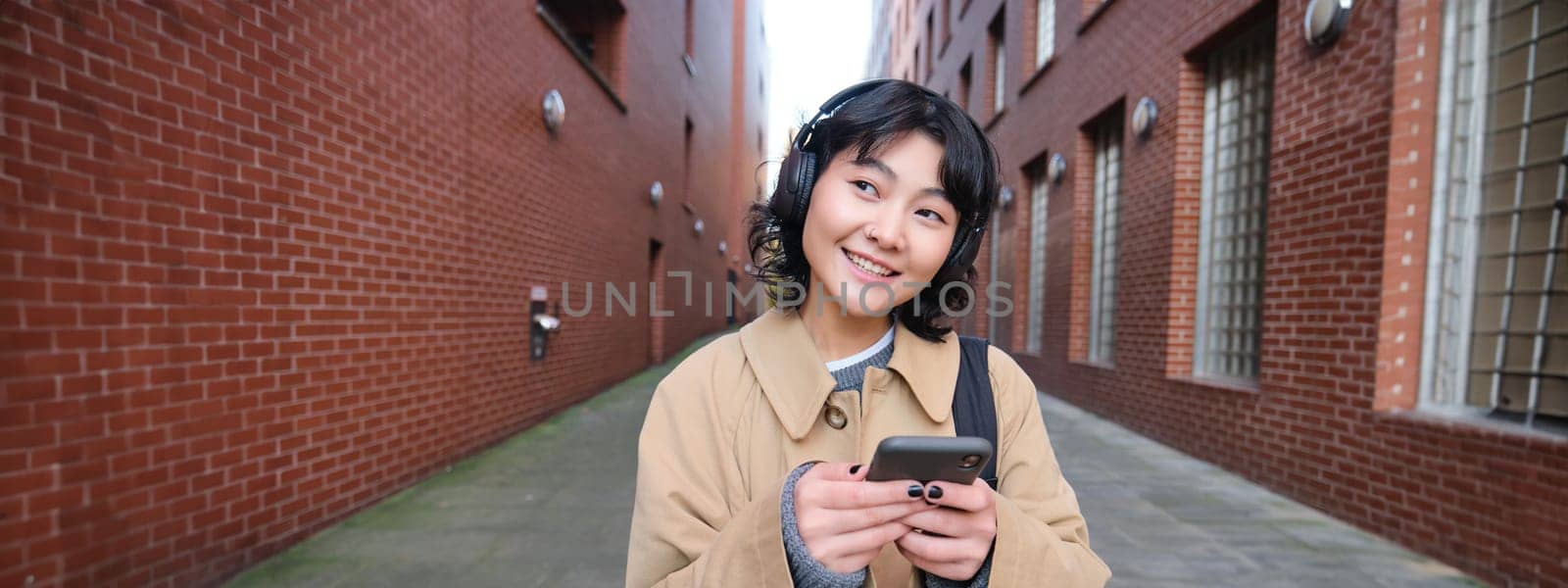 Portrait of young korean woman walking around city with backpack and headphones, listens music, looks at smartphone, uses phone application on streets by Benzoix