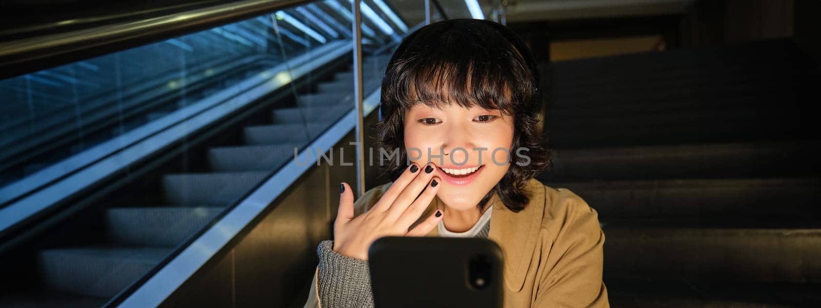 Korean girl looks surprised and happy at her phone screen, smiles amazed, reads good news, sits on staircase near escalator in mall, listens music in headphones.