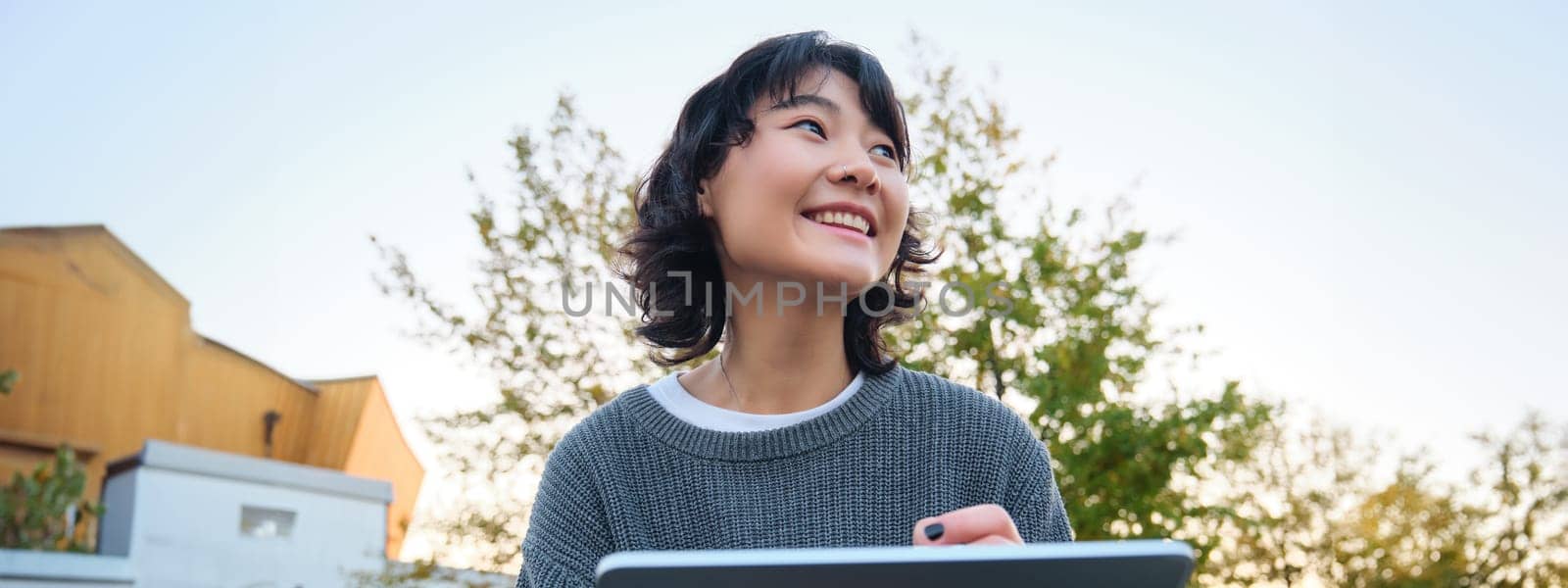 Close up portrait of young korean girl sits outdoors in park, holds her digital tablet and graphic pen, draws scatches, gets inspiration from nature for art, smiles happily by Benzoix