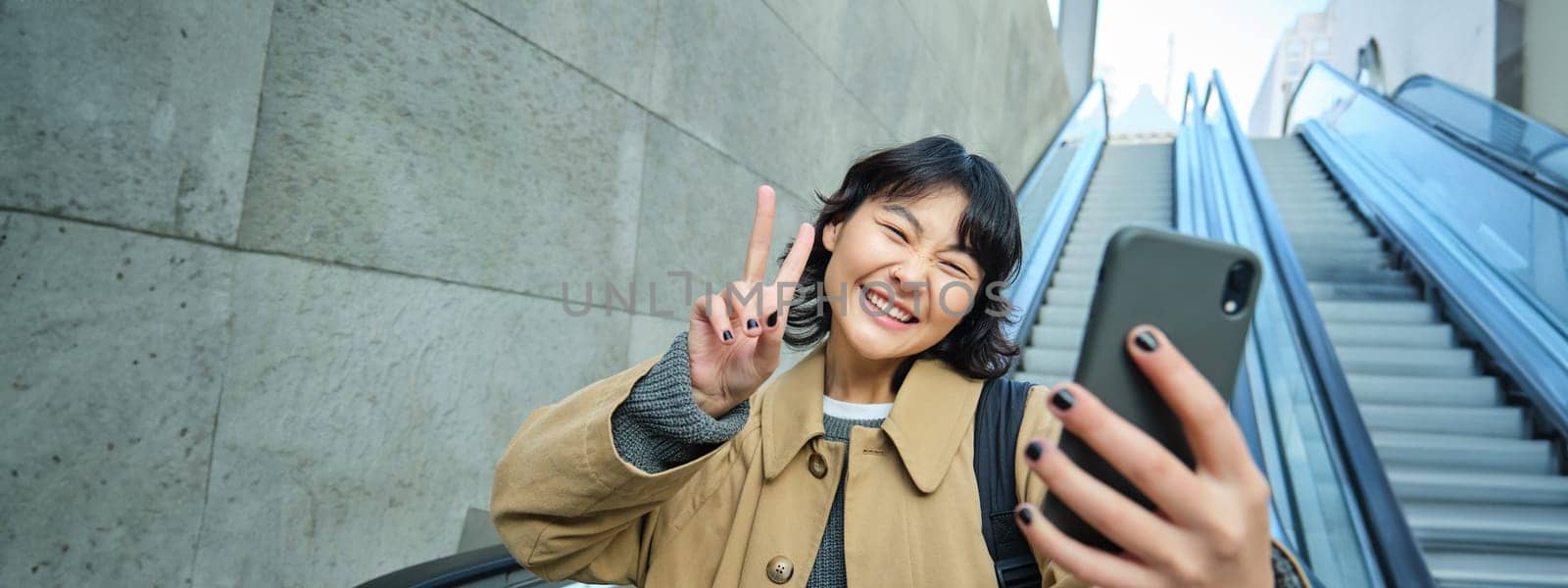 Happy triumphing asian girl, looks amazed and joyful, holds smartphone, goes down escalator with pleased face, wins, hears good news by Benzoix