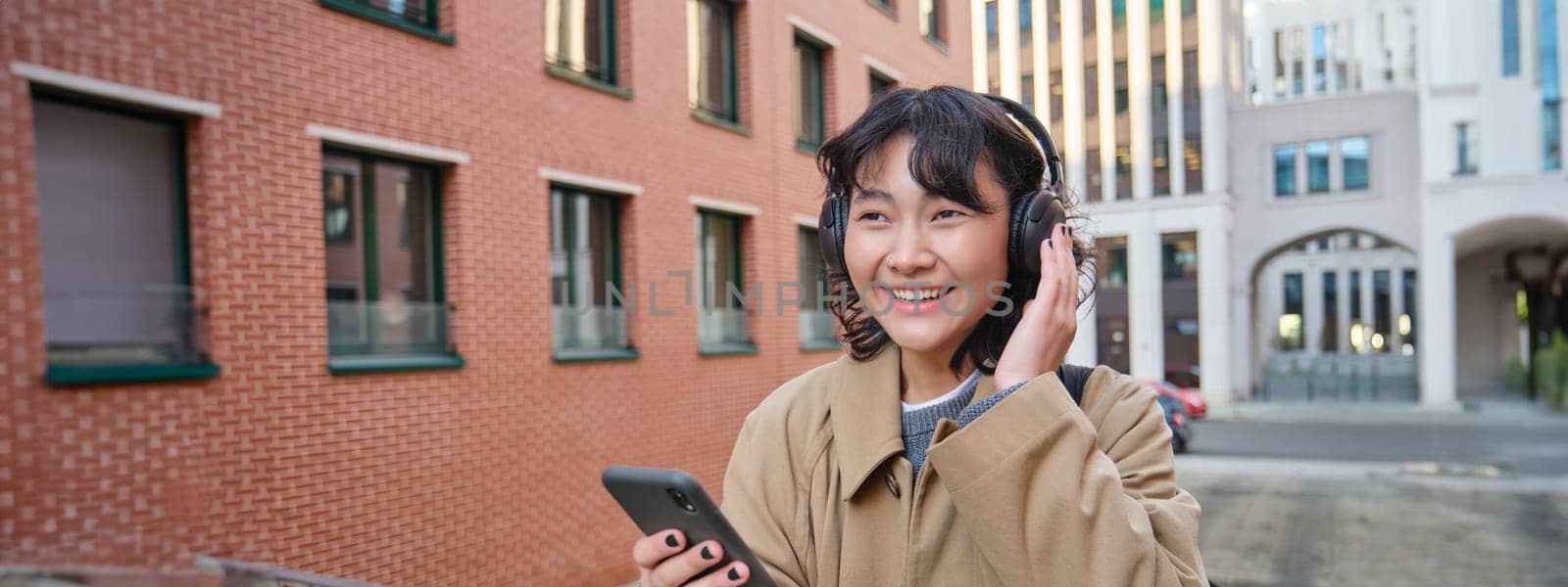 Young happy girl tourist, listens music in headphones, drinks takeaway and checks mobile phone, stands on street and smiles by Benzoix