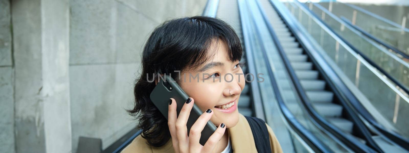 Headshot of smiling korean woman with smartphone, makes a phone call, goes down escalator in city, commutes to university by Benzoix