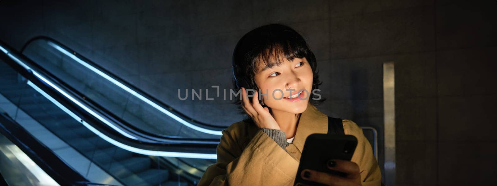 Cute young student, girl in headphones, commuting, standing near escalator, holding smartphone, listening music or podcast while travelling in city by Benzoix