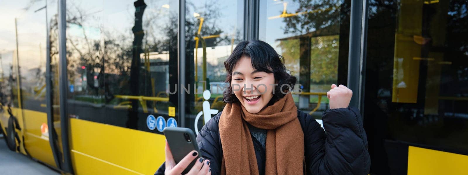 Portrait of cheerful asian girl talks on mobile phone, video chats, looks amazed at smartphone camera, stands on bus stop.