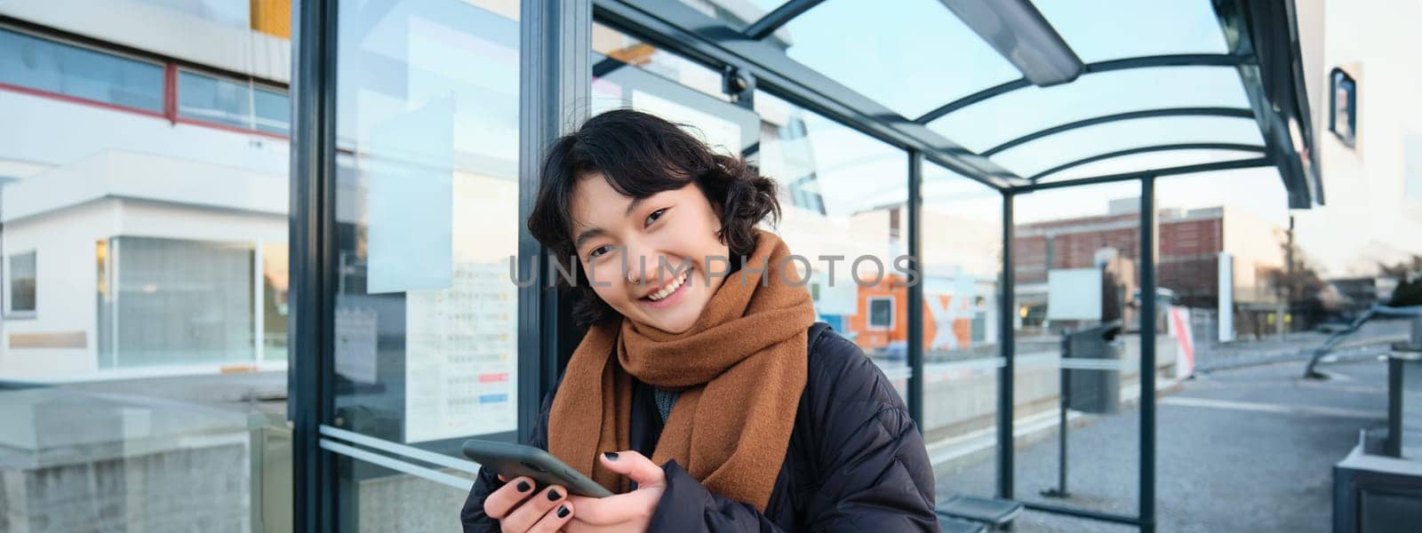 Cute smiling asian girl standing on bus stop, holding smartphone, wearing winter jacket and scarf. Woman commuting to work or university via public transport, stands on road by Benzoix