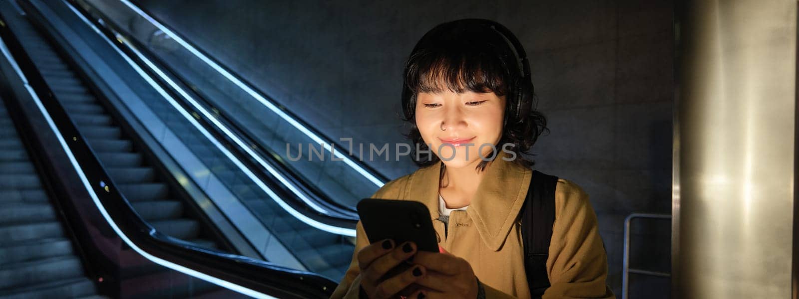 Smiling korean woman in headphones, reading message on smartphone, using mobile phone application, watching video or listens to music in public place by Benzoix