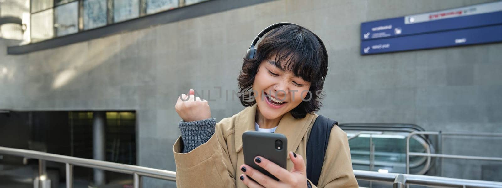 Cheerful Korean girl wins, celebrates victory, looks at smartphone app and triumphs, express joy and excitement, stands in headphones on empty street.