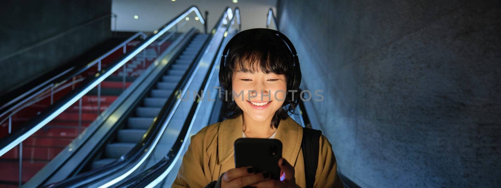 Beautiful young asian woman, going down an escalator, using smartphone, listening music in headphones while commuting to work on public transport by Benzoix