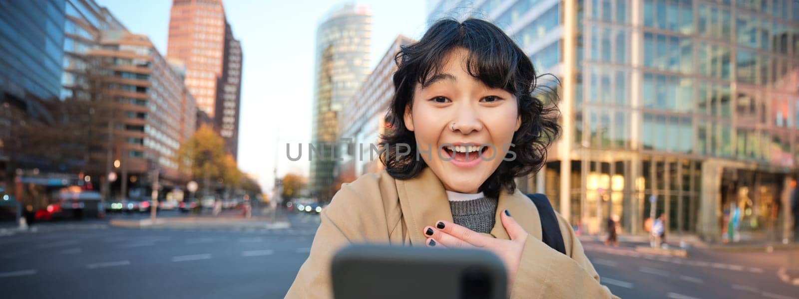 Portrait of asian girl looks surprised at smartphone screen, video chats and hears amazing news, looks impressed, stands on sunny street of city centre.