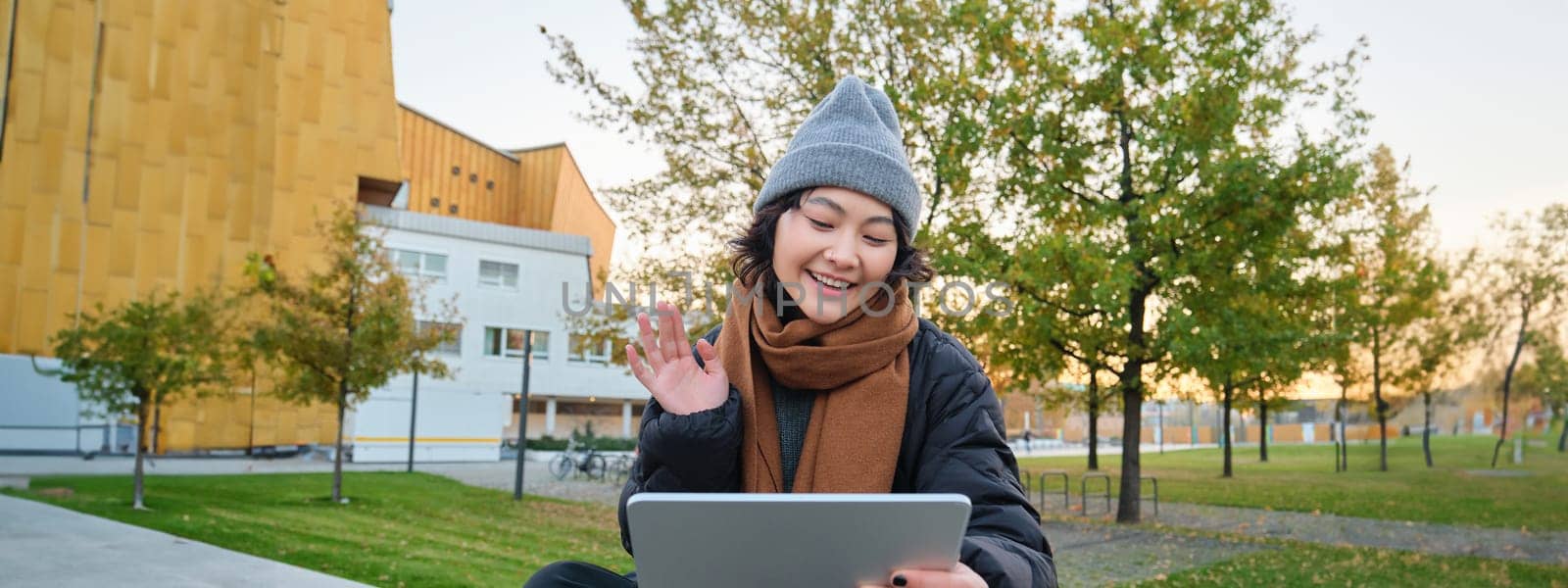 Portrait of young asian girl sits in warm clothes in park, waves hand at tablet, video chats outdoors, says hello while on call by Benzoix