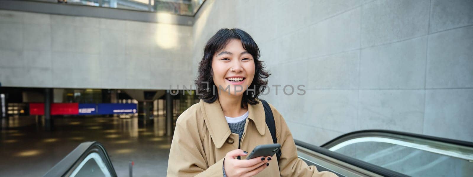 Technology and people concept. Smiling brunette woman travels around city, holds smartphone and laughs, uses mobile phone on her way home, stands on escalator.