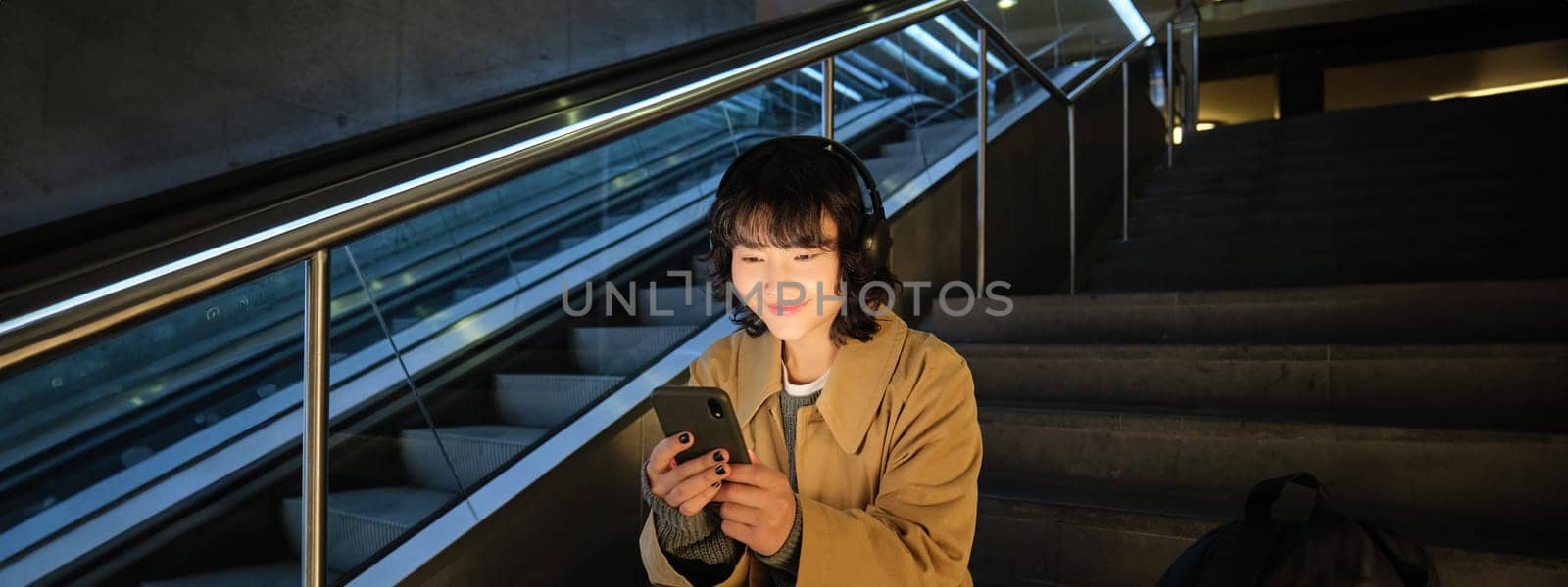 Technology. Portrait of asian girl sits on stairs near escalator, listens music in headphones and uses social media, reads, watches video in public place.