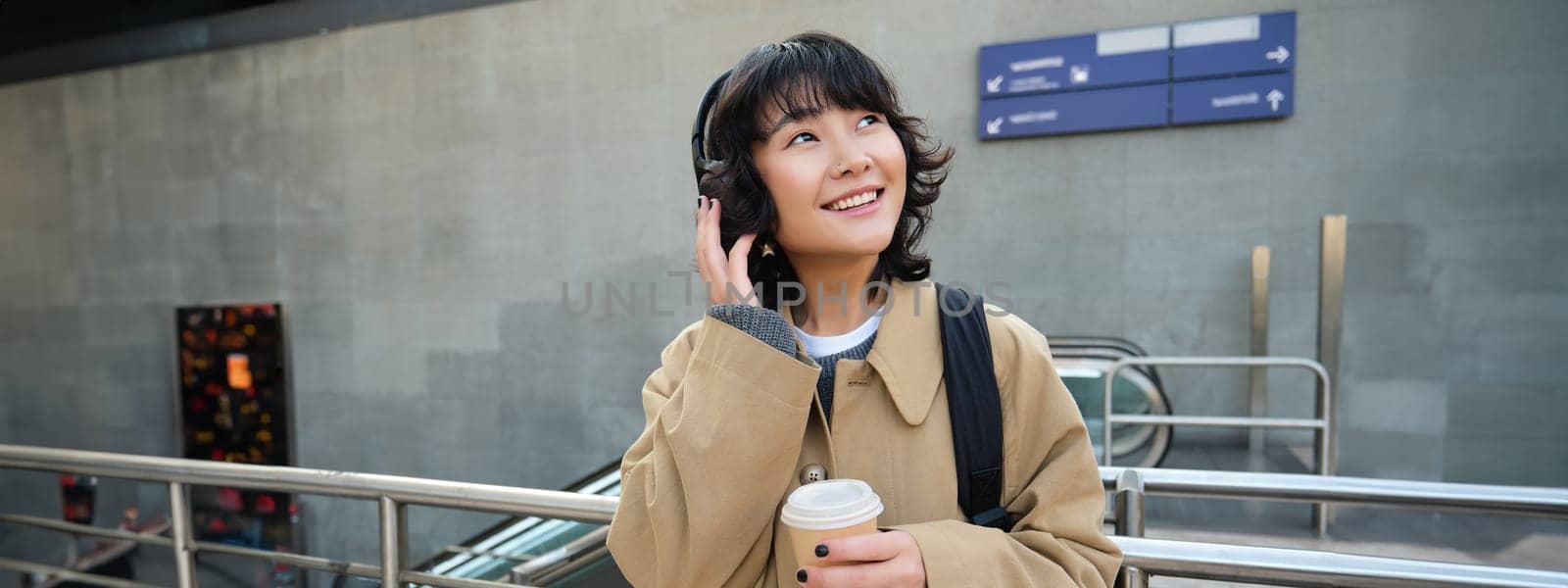 Portrait of young student, girl in headphones, drinks coffee, stands on street with backpack, commutes to university or college, smiles happily.