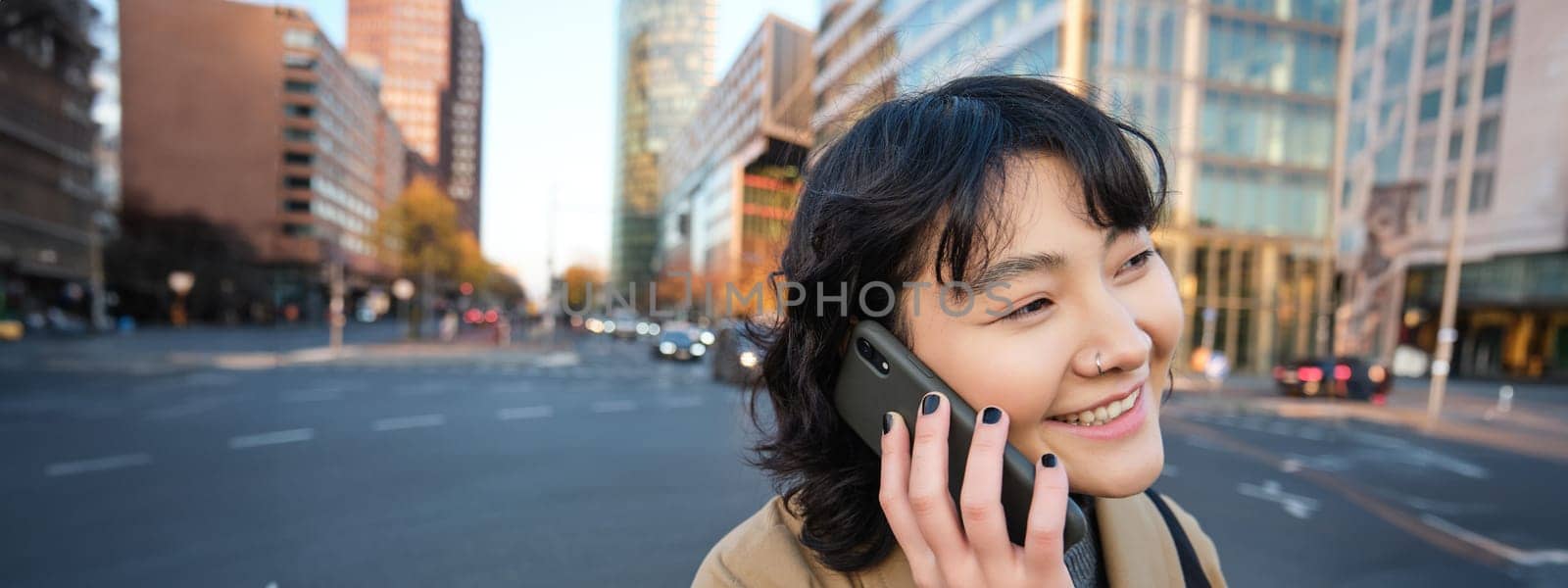 Smiling asian girl makes a phone call, stands on an empty street, calling someone on telephone, waiting for friend in city, going to a meeting by Benzoix