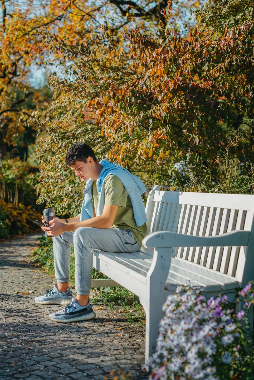 A Teenager Sits On A Bench In The Autumn Park Drinks Coffee From A Thermo Mug And Looks Into A Phone. Portrait Of Handsome Cheerful Guy Sitting On Bench Fresh Air Using Device Browsing Media Smm Drinking Latte Urban Outside Outdoor by Andrii_Ko