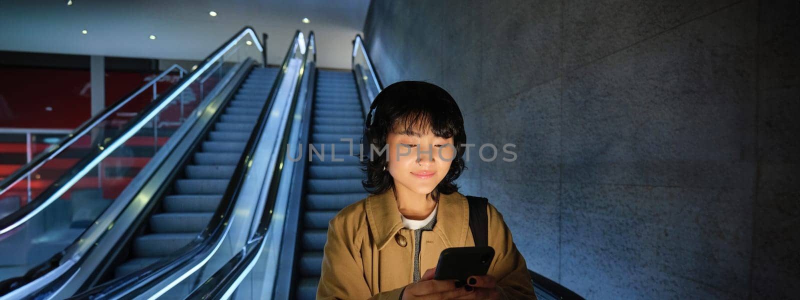 Cute young woman going down the escalator to the tube, using subway metro to commute to work or university, standing with smartphone by Benzoix