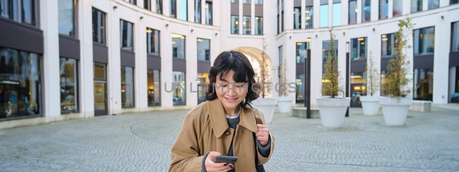 Technology, application and cellular concept. Smiling asia woman uses mobile phone, holds smartphone and reads text message, walks on streets of city centre.