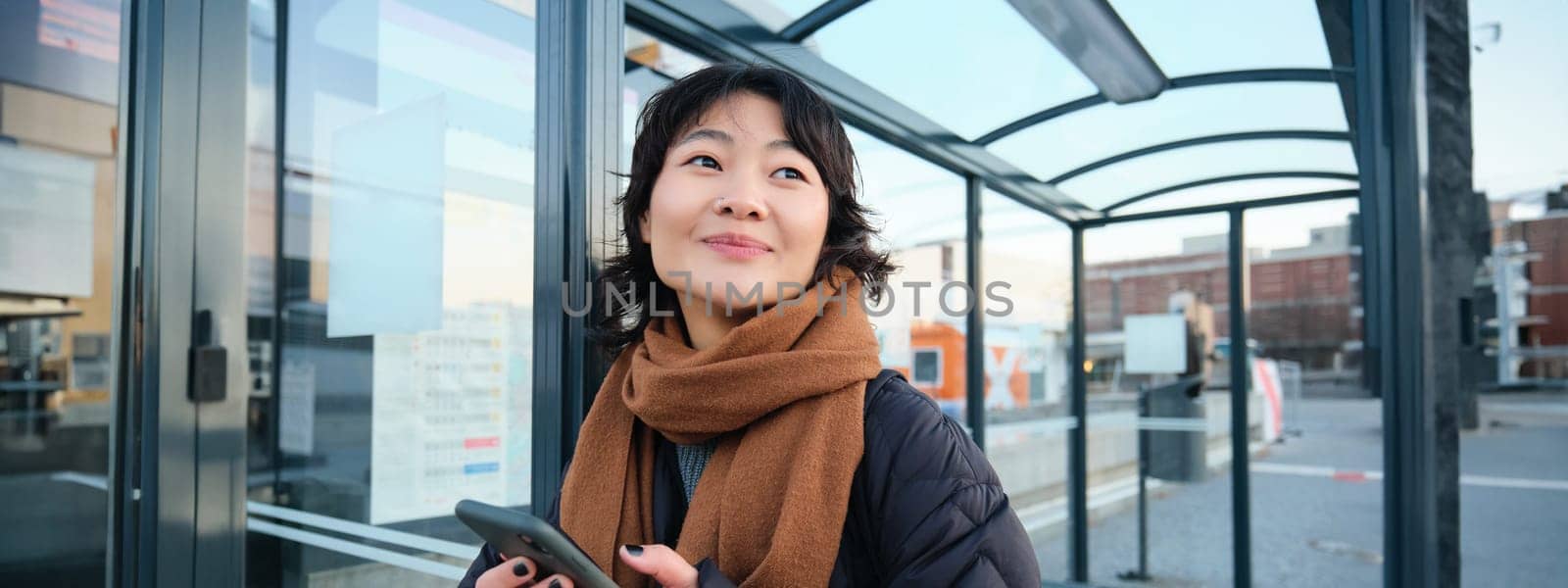 Beautiful Japanese girl, student standing on bus stop in winter jacket, holding smartphone, using application to track her public transport, ordering a ride.