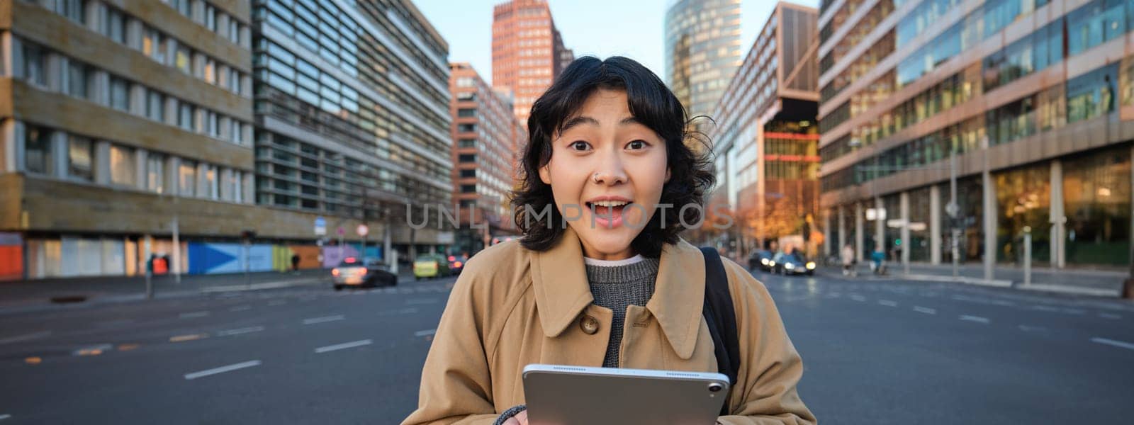 Portrait of korean girl looks surprised, found out something amazing, holding digital tablet with joyful face, stands on street of city centre by Benzoix