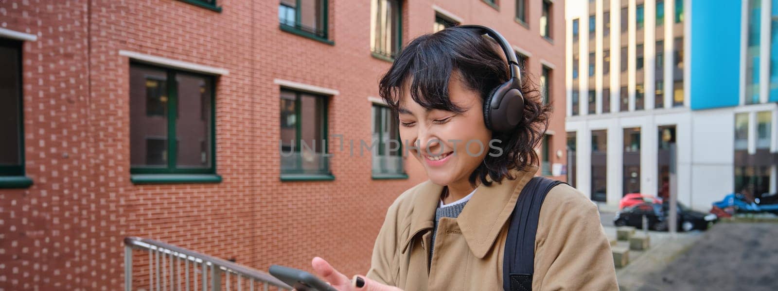 Happy korean girl looks at mobile phone and listens music in headphones, stands on street in city centre, reads text message on smartphone.