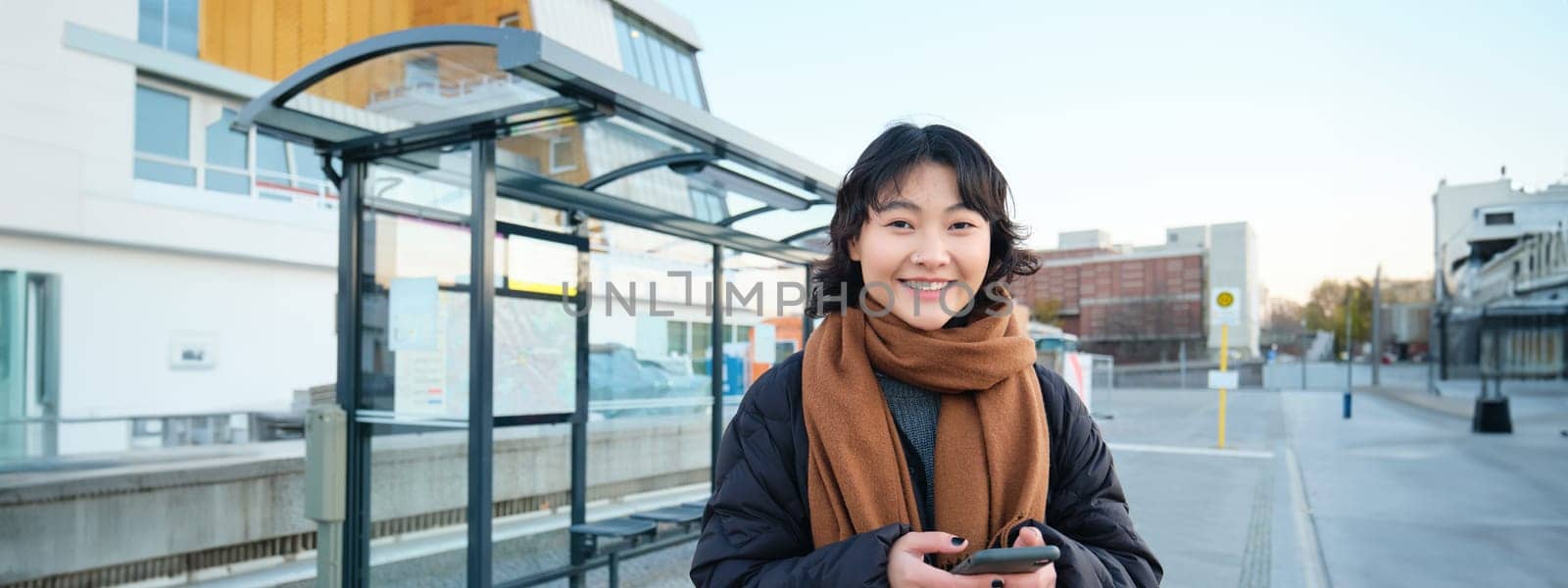 Happy smiling korean girl, using mobile phone, standing on bus stop with smartphone, looking at departure schedule on application, posing in winter clothes by Benzoix