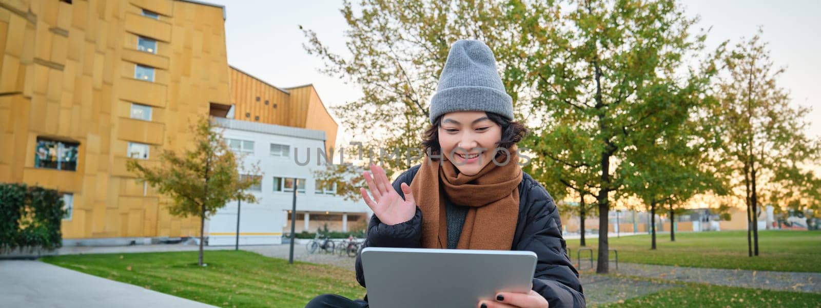 Portrait of young asian girl sits in warm clothes in park, waves hand at tablet, video chats outdoors, says hello while on call.