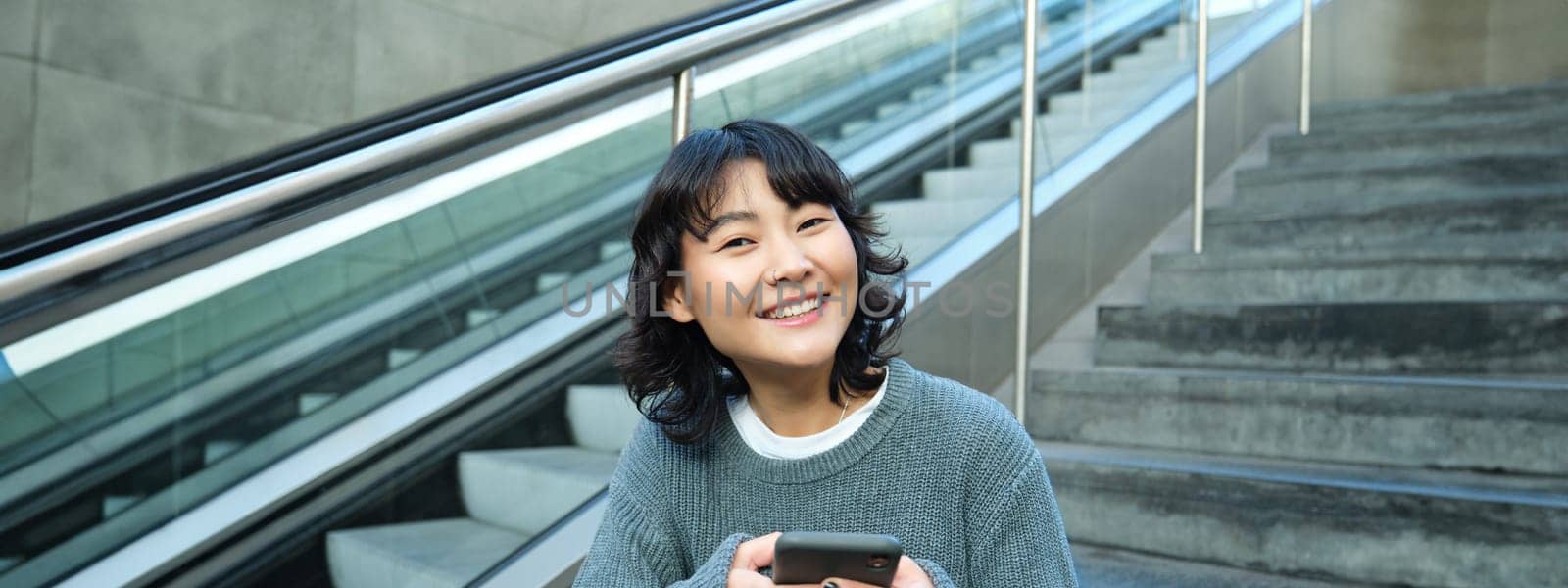 Portrait of brunette young woman, student sits on stairs in public place, looks at smartphone, reads text message and smiles.