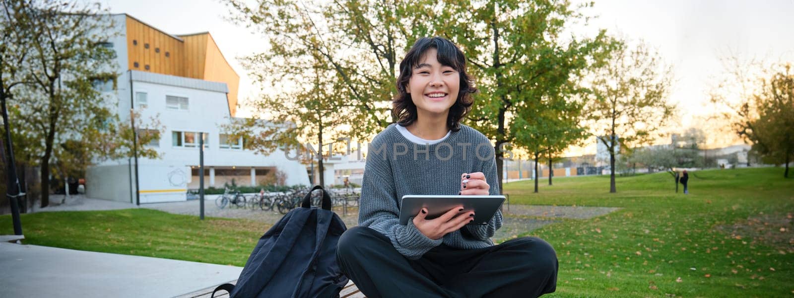 Young asian girl, student sits on bench, graphic designer with pencil and digital tablet draws scatches, does her homework outdoors.